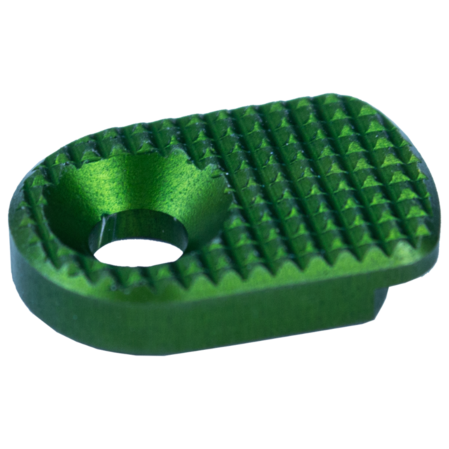 Close-up of the MAGAZINE CATCH PADDLE SHADOW 2 GREEN, a small metallic object with a textured surface, featuring a round hole in the center and a grid-like pattern for grip or aesthetics.