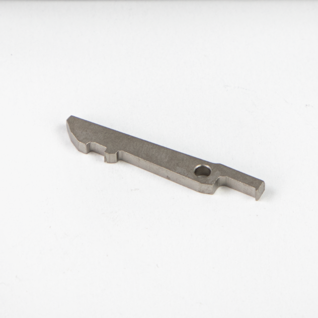The FIRING PIN BLOCK BREN 2 is a flat, thin metal piece with a straight edge on one side and a notched, angled edge on the other. It includes a small circular hole near the end of the angled side and is displayed on a plain white surface.