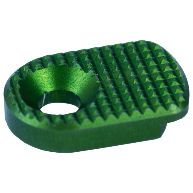 Close-up of the MAGAZINE CATCH PADDLE SHADOW 2 GREEN, a small metallic object with a textured surface, featuring a round hole in the center and a grid-like pattern for grip or aesthetics.