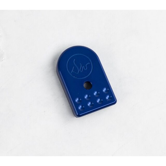 The DWX MAGAZINE BASE PAD BLUE is a blue plastic item with a rounded top and flat bottom, featuring a central hole and an embossed script logo. It also includes six small raised dots arranged in a line near the bottom edge, set against a plain white backg