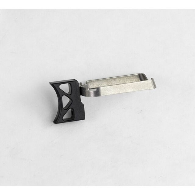 On a plain white surface lies the DWX FULL SIZE TRIGGER ASM, CURVE K, BLAC—a small component crafted from metal and plastic. It features a dark curved section with triangular cutouts on one end and a flat metallic part with a clip on the other end.